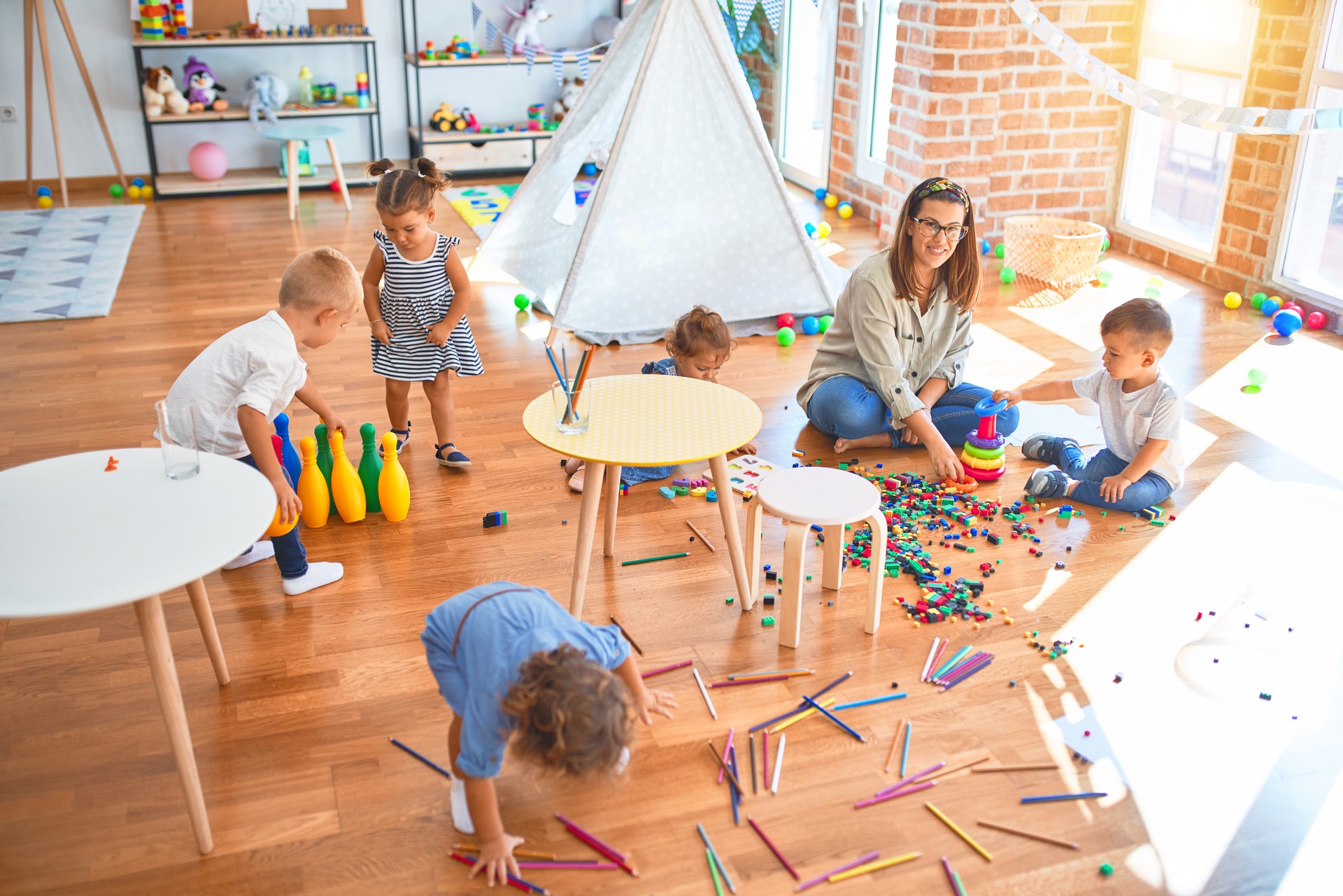 Beautiful teacher and group of toddlers playing around lots of toys at kindergarten